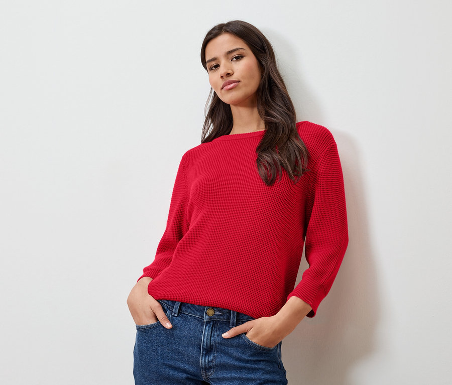 Nő piros, finom kötött pulóverben és farmerban, fehér fal előtt pózol.