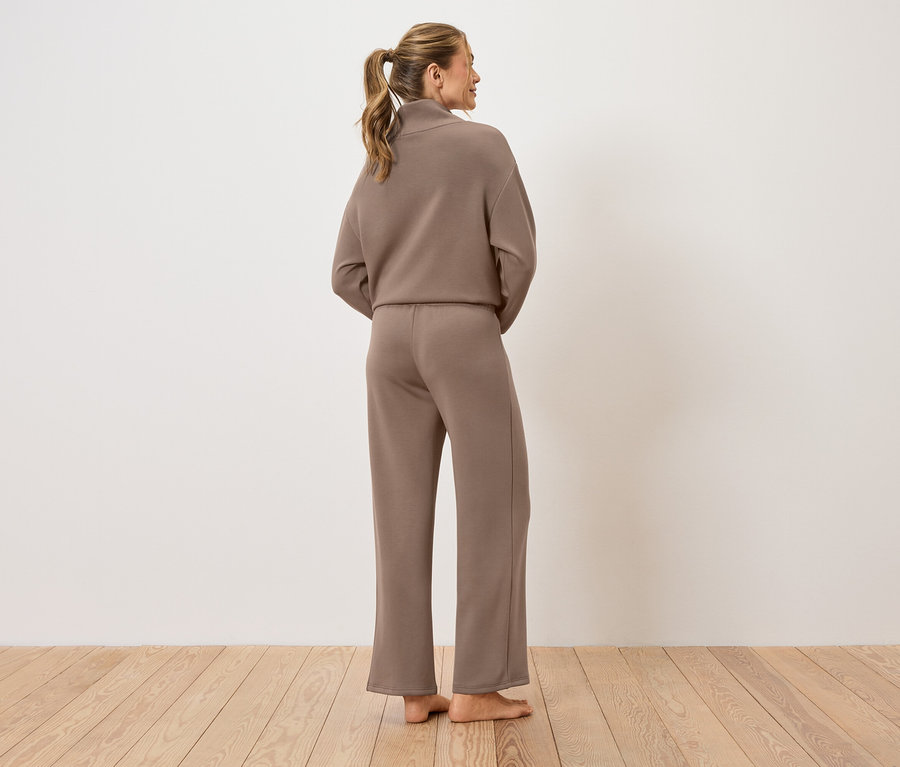 Egy nő áll háttal a kamerának, barna nadrágkosztümöt visel.