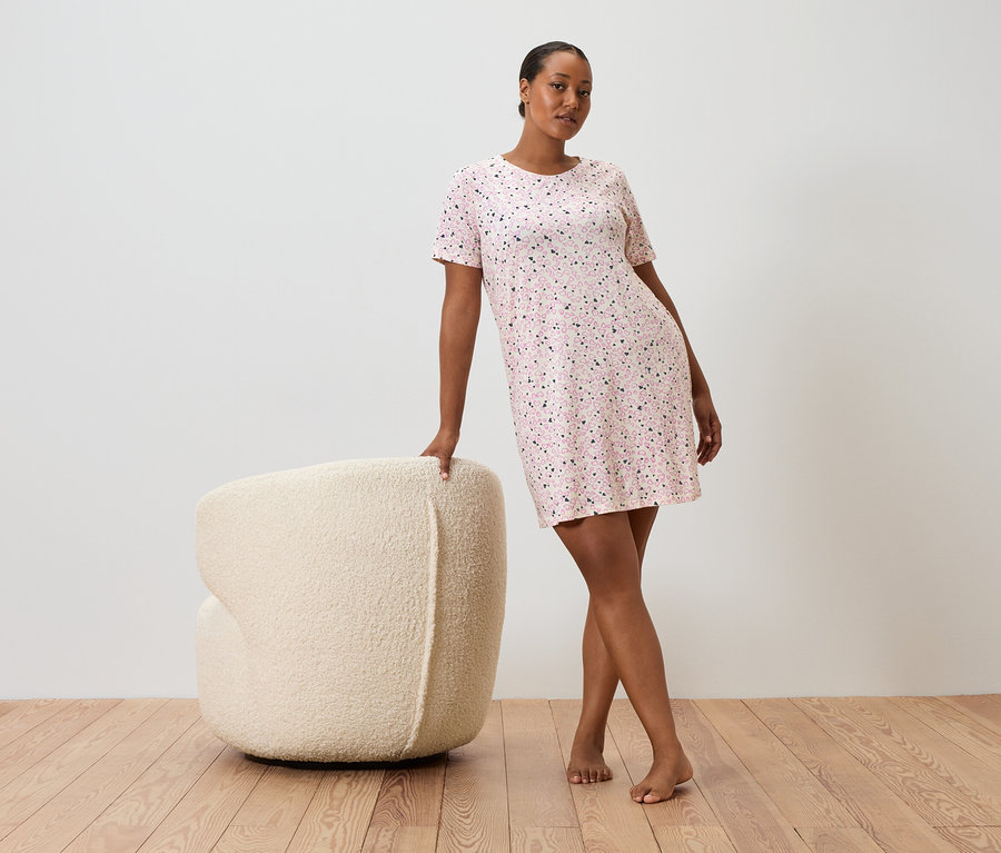 Egy nő egy krémszínű karosszéknek támaszkodik, rövid ujjú, rózsaszín pöttyös ruhát visel.