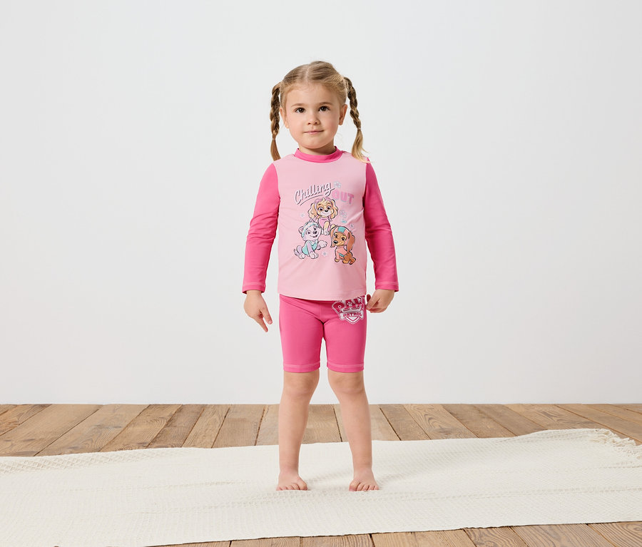 Egy kis lány fonott hajjal mezítláb áll egy krémszínű szőnyegen, rózsaszín Paw Patrol hosszú ujjú pólót és rövidnadrágot visel.