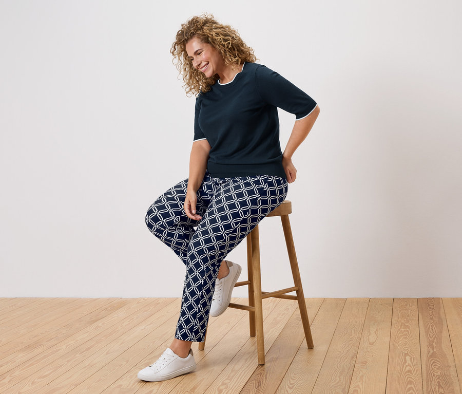 Göndör hajú nő ül egy széken, sötétkék felsőt és mintás nadrágot visel.