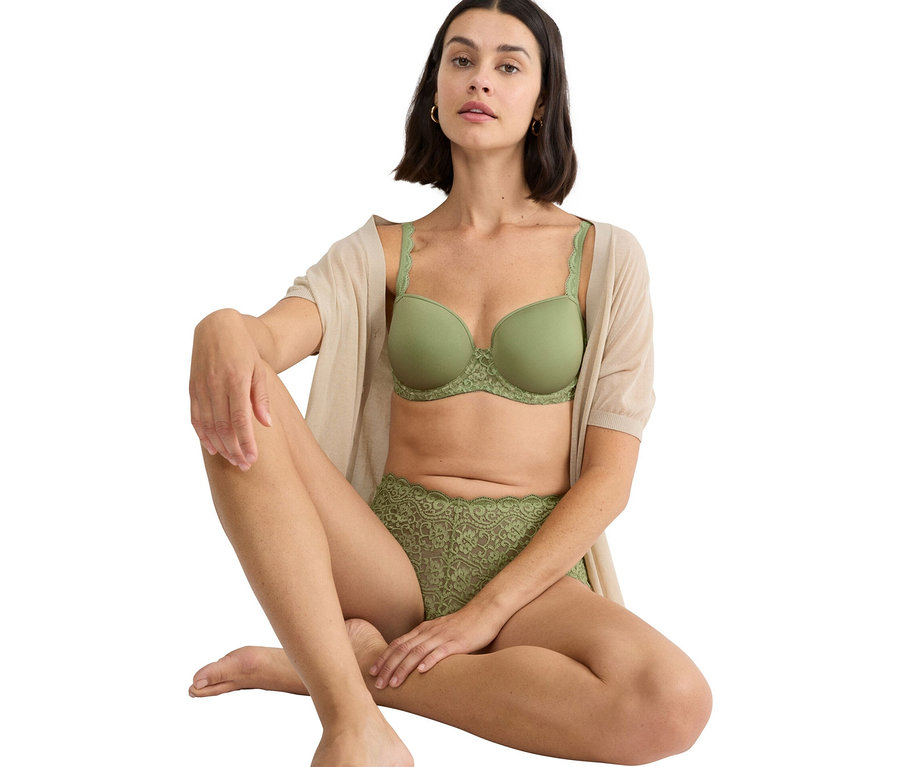 Nő ül keresztbe tett lábakkal, zöld melltartót és zöld csipkés bugyit visel, valamint egy bézs kardigánt.