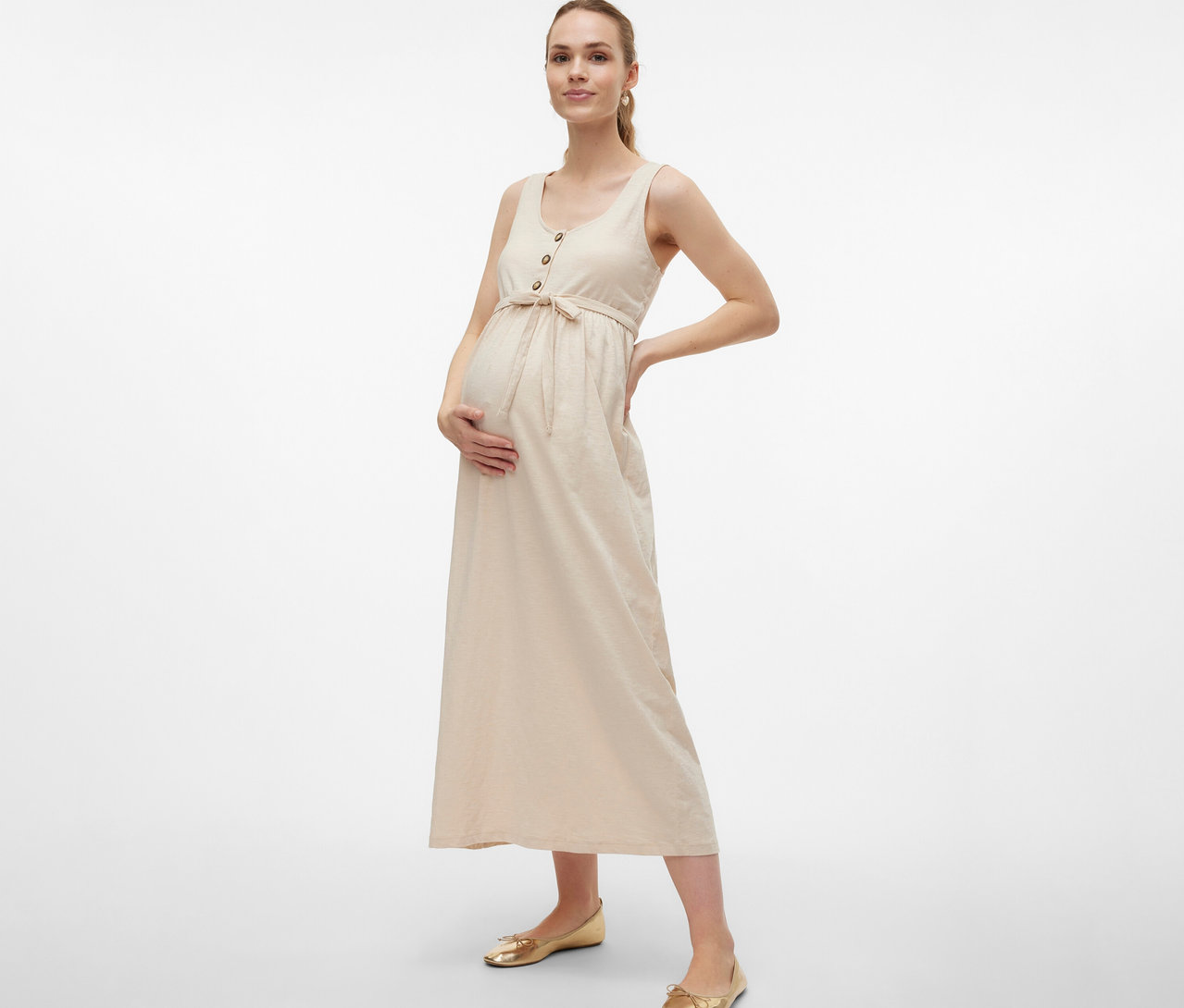 Egy várandós nő a MAMALICIOUS »MLEVI LIA« kismamaruhában, övvel és gombokkal, a kezét a hasán és a csípőjén tartva pózol.