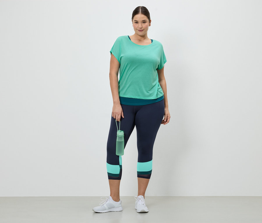 Egy nő sportruházatban, amely egy zöld pólóból, sötétkék leggingsből és fehér tornacipőből áll, egy zöld vizes palackot tart.
