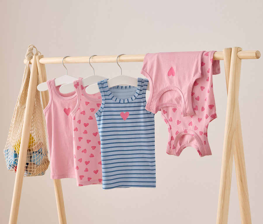 Egy fa rúdon rózsaszín és kék csíkos, szívecske mintás gyerek trikók lógnak.