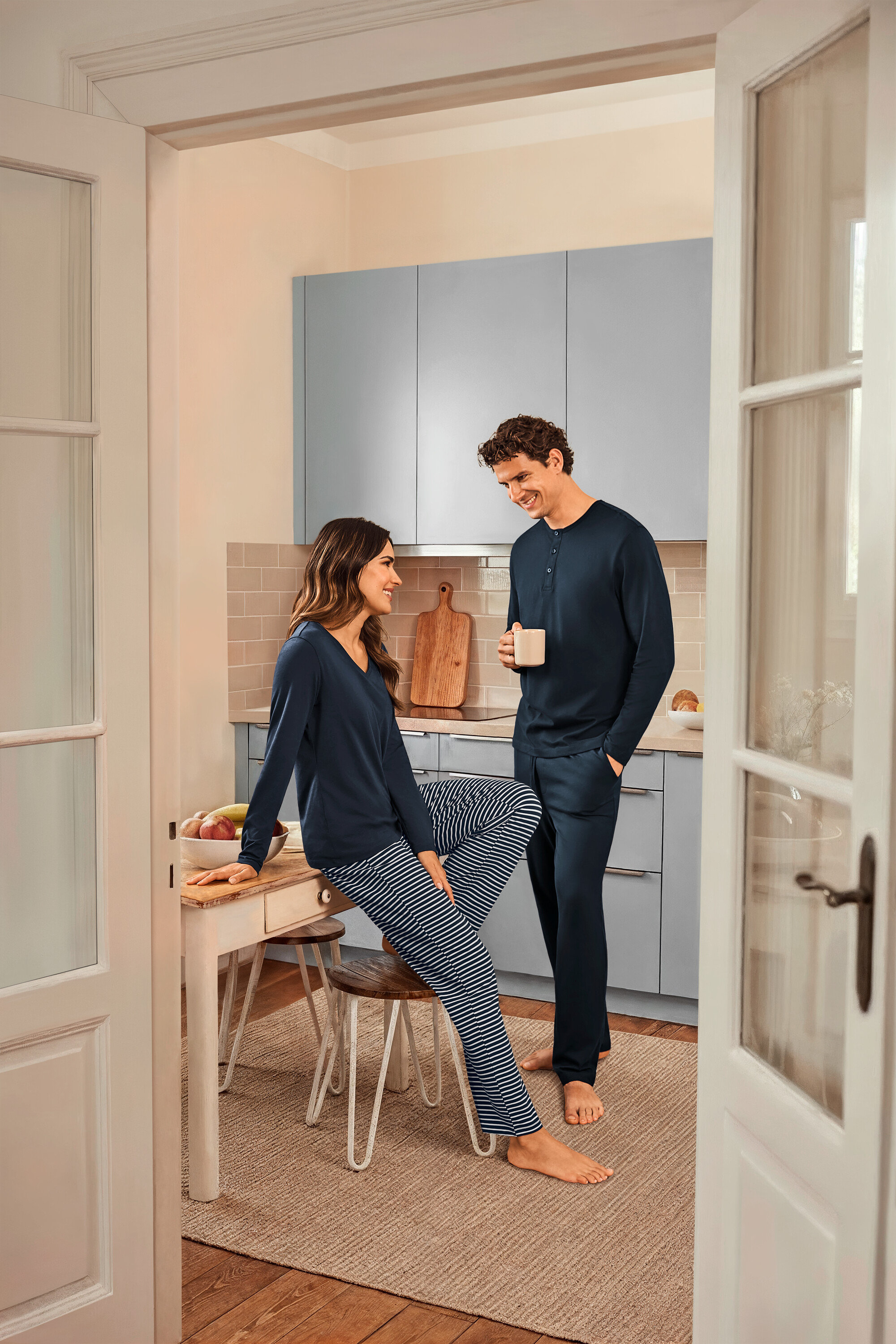 Egy konyhában egy nő ül egy asztalon sötétkék csíkos pizsamában és sötétkék felsőben, egy férfi pedig egy csészét tart a kezében és sötétkék pizsamát visel.