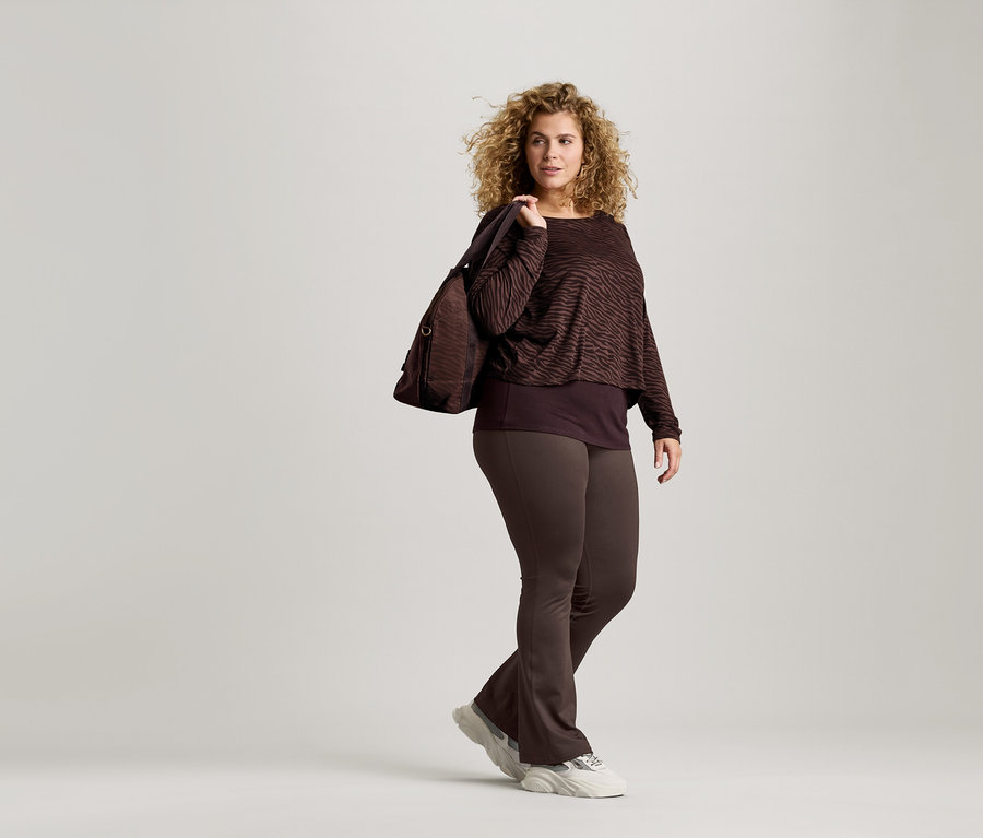 A göndör hajú nő barna, zebracsíkos felsőt, barna leggingst és edzőcipőt visel. Barna táska van a vállán.