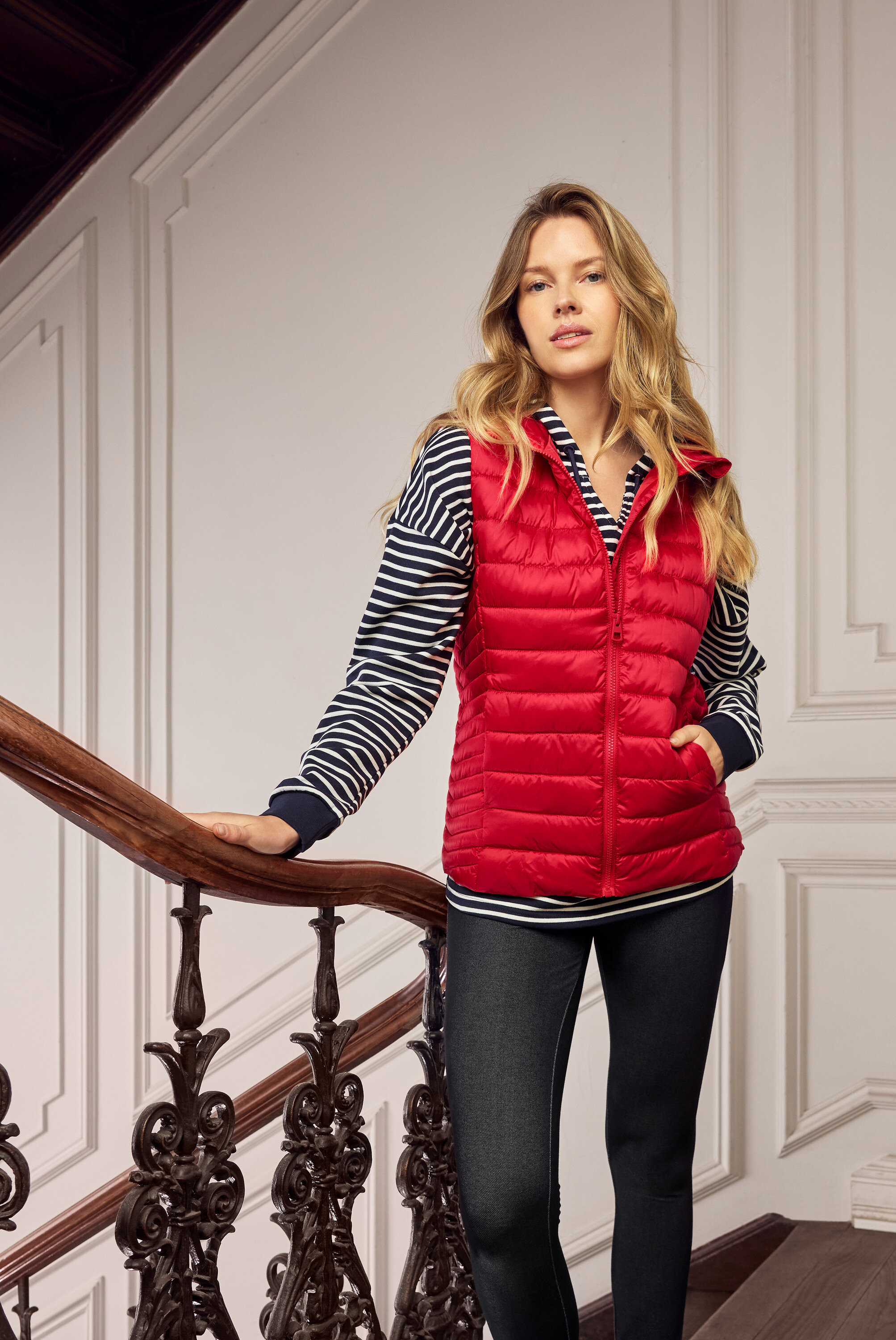 Szőke nő piros steppelt mellényben, kék-fehér csíkos felsőben és sötét farmerben egy lépcsőn.