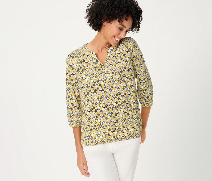 Mosolygó nő visel egy sárga mintás Olsen 3/4-es ujjú pólóblúzt és fehér nadrágot.