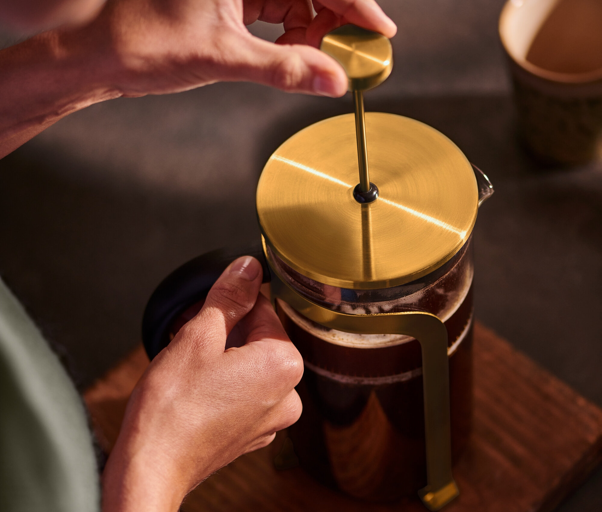 Egy személy egy arany színű, 800 ml-es, 6 csészés dugattyús kávéfőzőt nyom le egy fa alátéten.