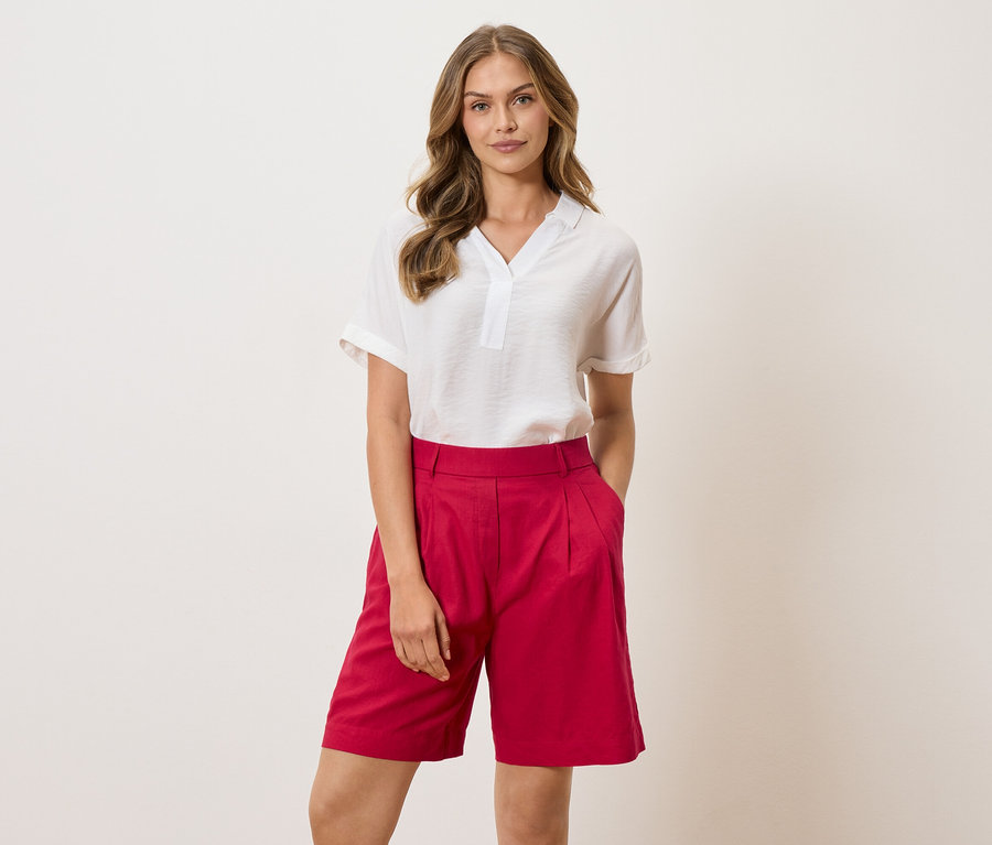 Nő fehér blúzban és piros rövidnadrágban áll világos háttér előtt.