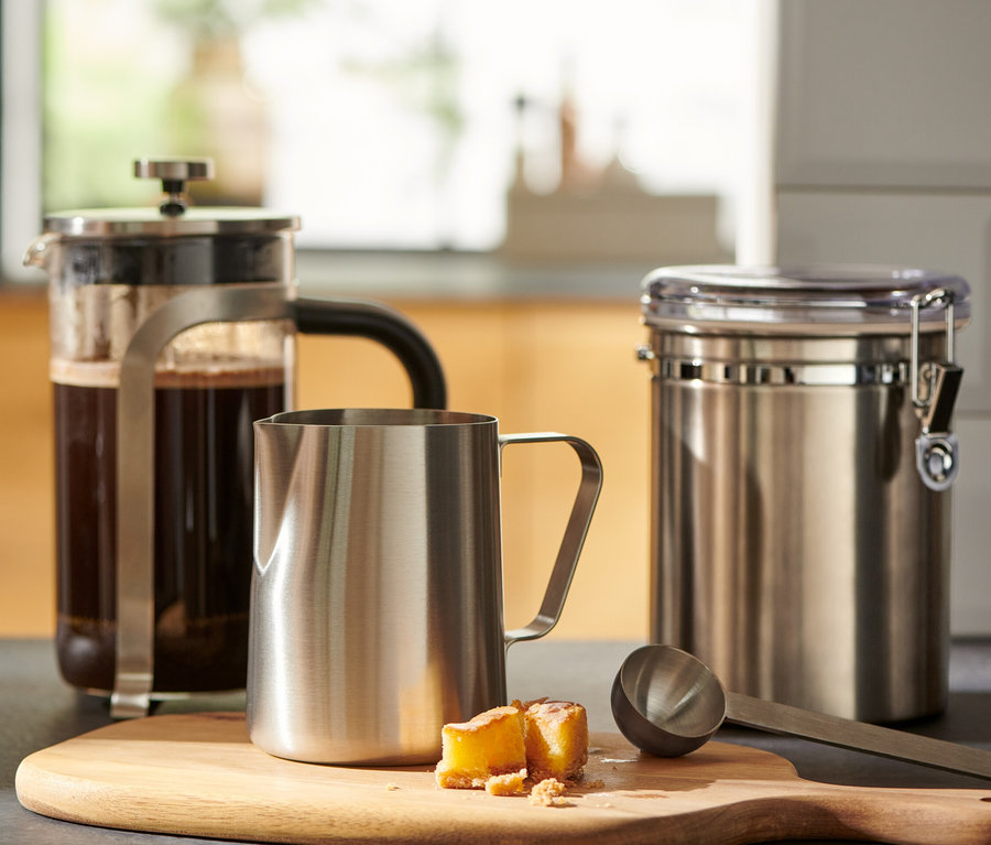 Egy pohár kávé egy french press mellett áll, és egy kis, nemesacélból készült tejeskancsó egy fa vágódeszkán.
