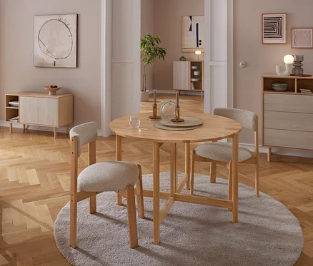 Egy étkezőhelyiség kerek fa asztallal és három világos kárpitozott székkel egy szürke szőnyegen. Az asztalon gyertyatartók és egy pohár található. A háttérben egy fa tálalószekrény és egy komód áll semleges falak előtt, absztrakt művészettel, mindez parkettán.