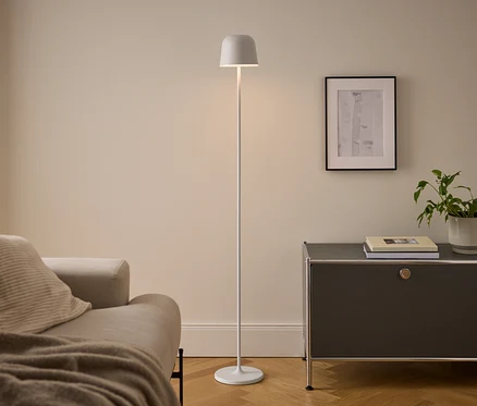 Modern nappali bézs kanapéval, fehér állólámpával és sötétszürke szekrénnyel. A megvilágított lámpa a kanapé és a szekrény között áll, utóbbin könyvek és egy cserepes növény van egy bekeretezett kép alatt.