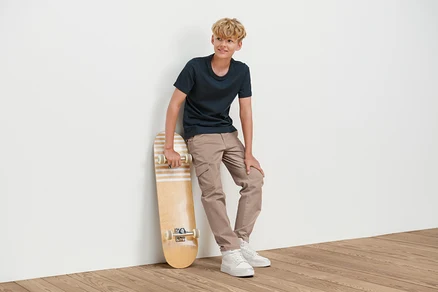 Ein Junge lehnt an einer Wand und hält ein Skateboard. Er trägt eine Kinder-Cargohose.