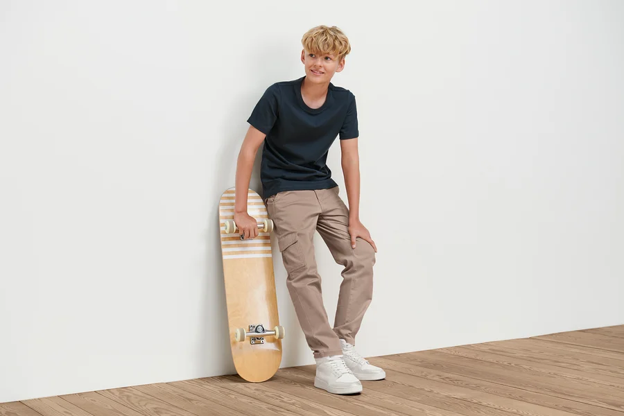 Ein Junge lehnt an einer Wand und hält ein Skateboard. Er trägt eine Kinder-Cargohose.