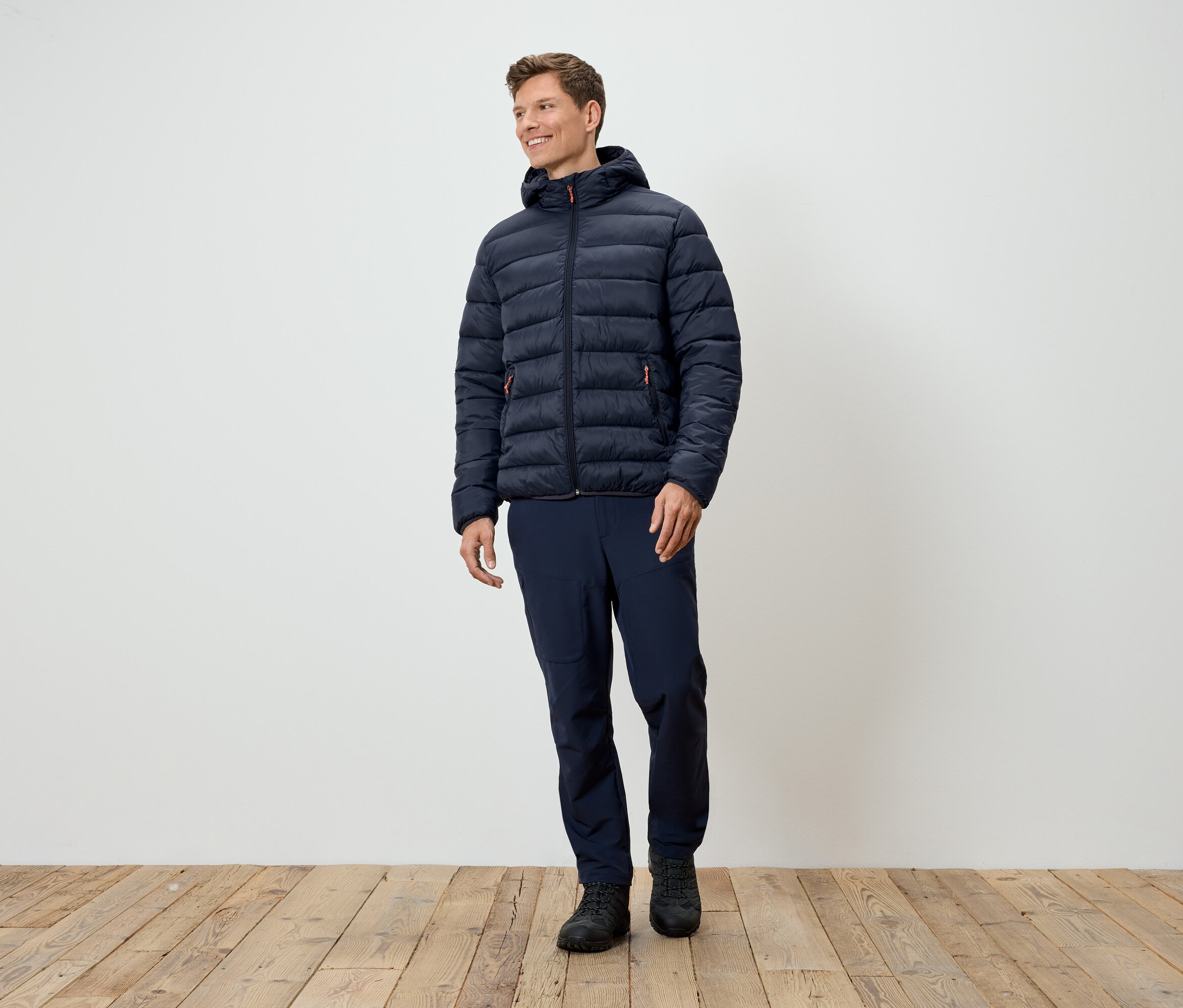 Egy férfi áll sötétkék steppelt kabátban, sötétkék nadrágban és fekete cipőben.