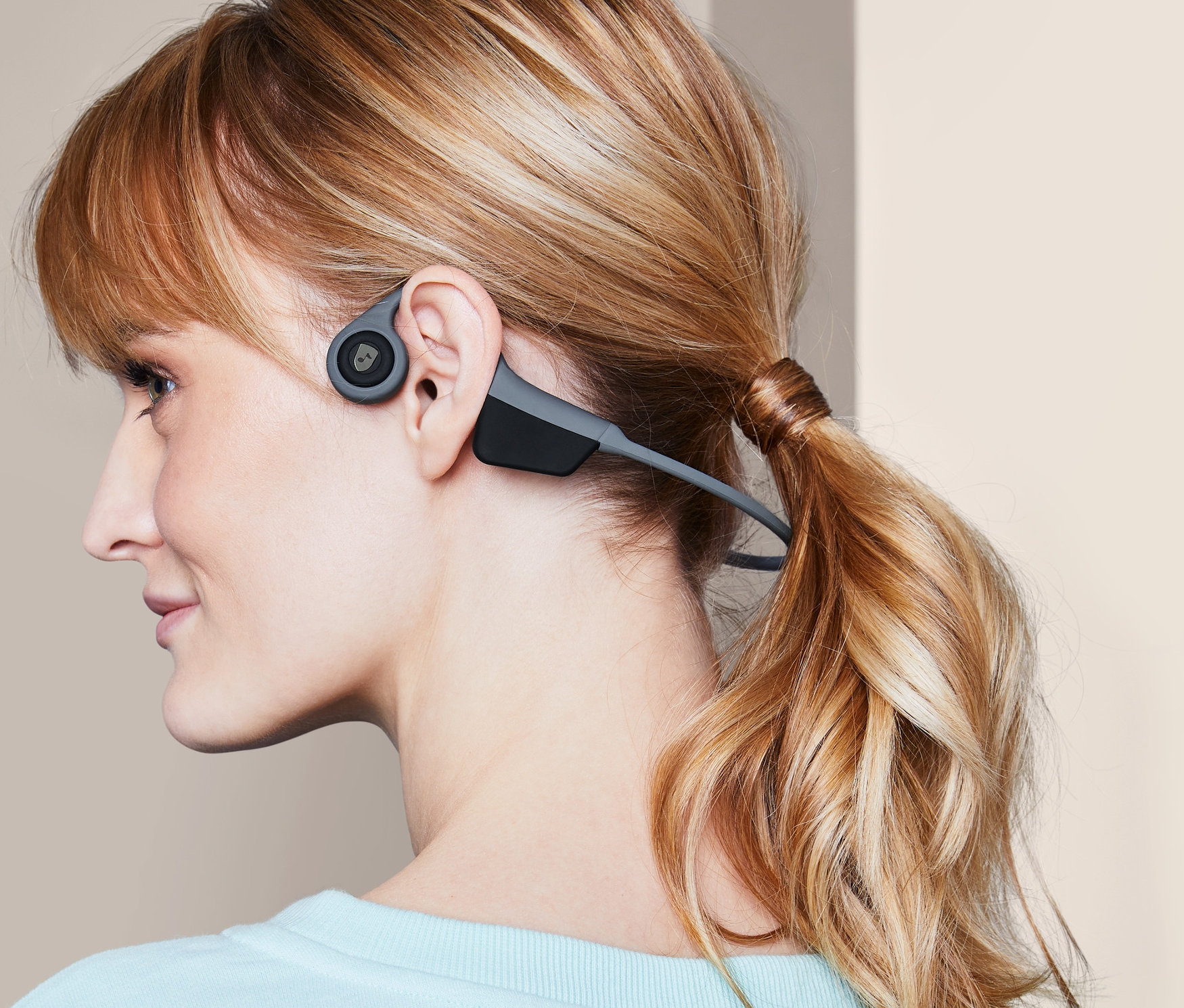 Egy szőke hajú, lófarokba kötött nő profilja, aki szürke, csontrezgéses fejhallgatót visel.