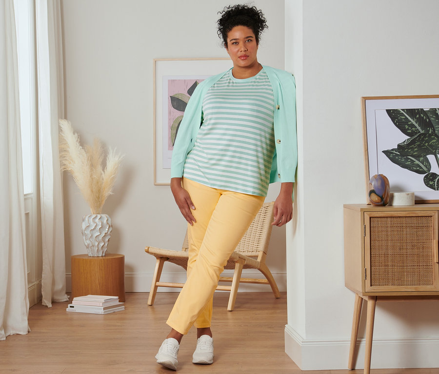 Egy falnak támaszkodó nőn csíkos, menta póló, menta kardigán, sárga slim fit farmer és krémszínű velúrbőr sneaker cipő van.