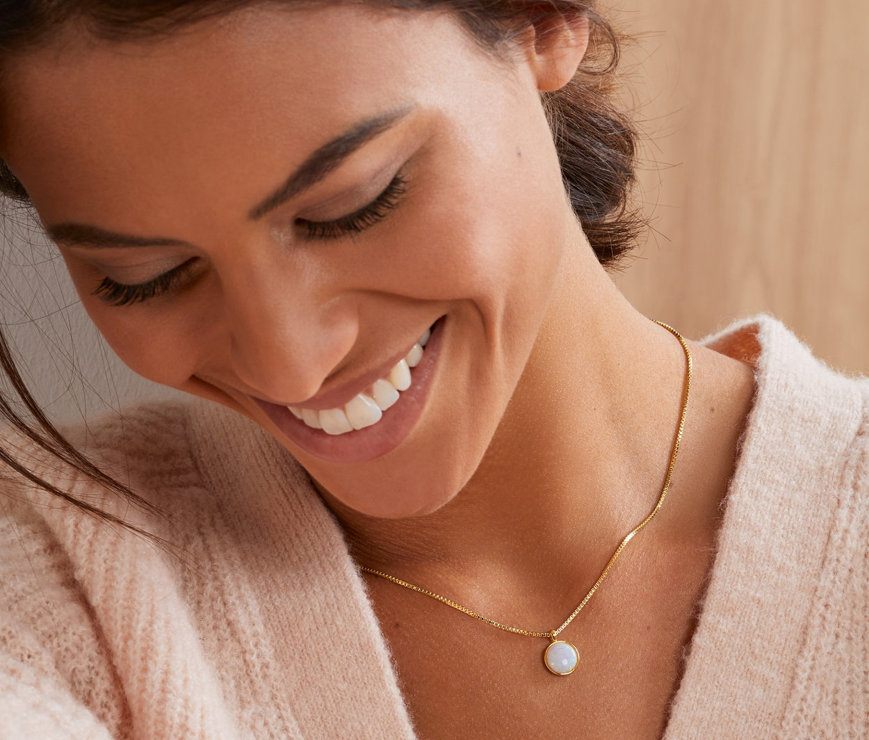 Mosolygó nő megérinti a haját, és egy arannyal futtatott, szintetikus opál medállal ellátott női nyakláncot visel.