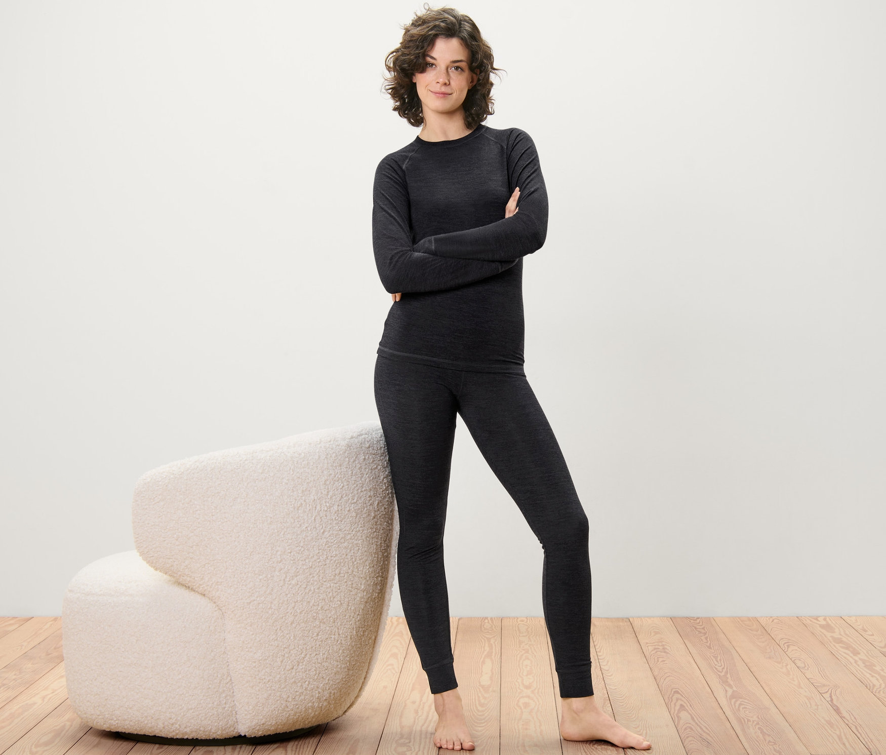 Nő pózol RWS-tanúsítvánnyal rendelkező gyapjúból készült hosszú alsóban és felsőben, fapadlón, egy krémszínű fotel mellett.