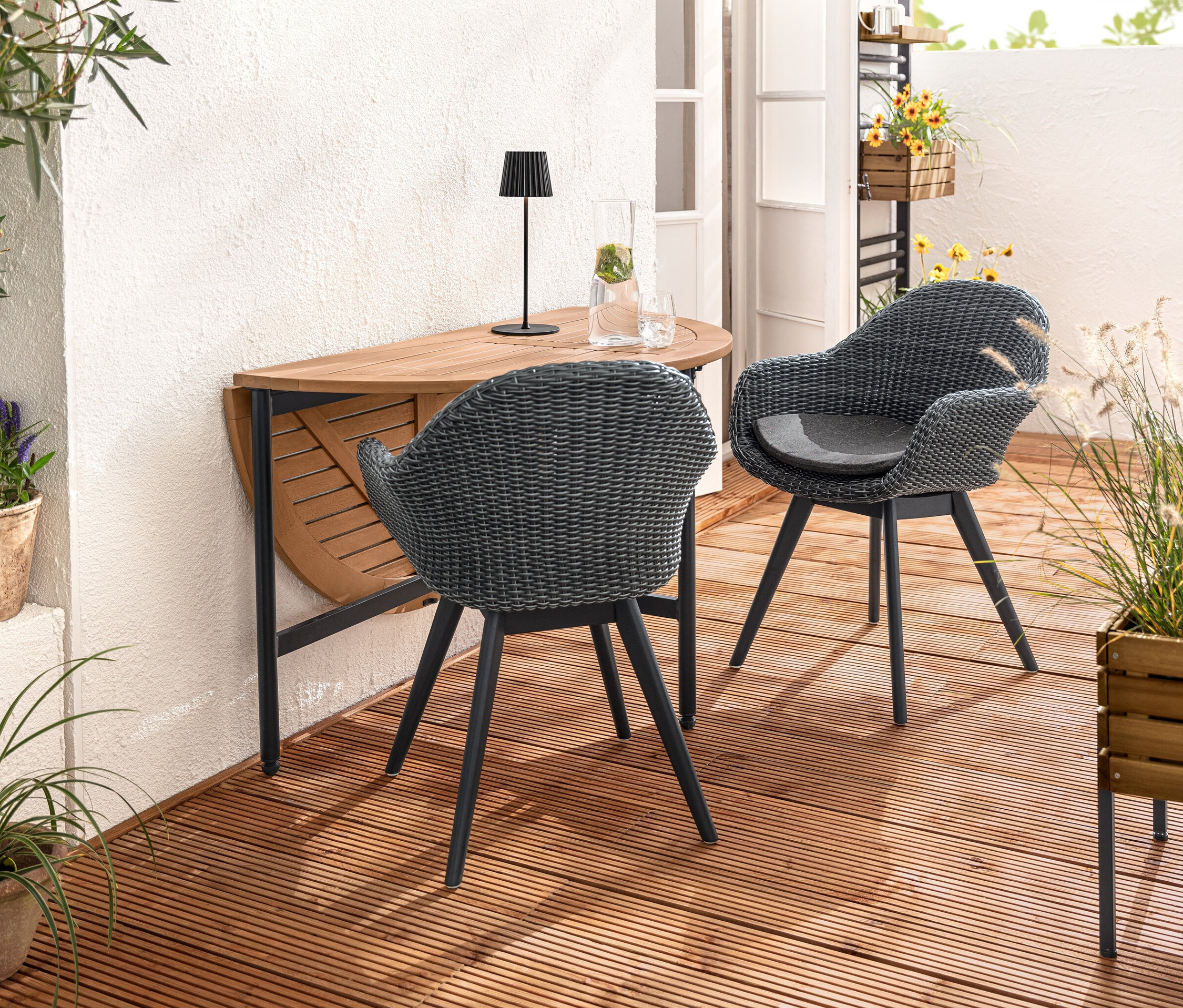 Két kagylófotel fonott polirattannal egy »Elin« lehajtható asztal mellett áll. Az asztalon LED-es akkumulátoros kültéri asztali lámpa (fekete) van. Elöl egy »Elin« ültetőláda látható.