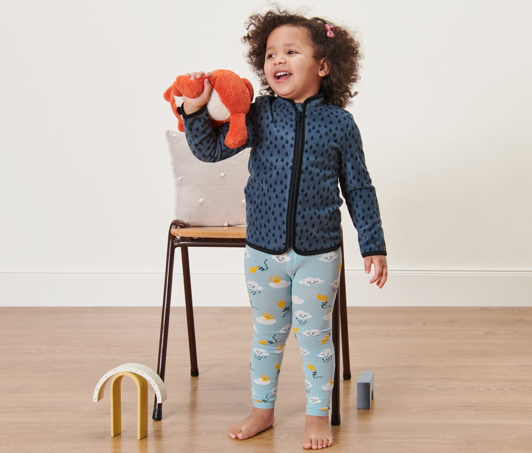 Mosolygó kislány plüss rókát tart. Felhős kék leggingset és kék pulóvert visel.