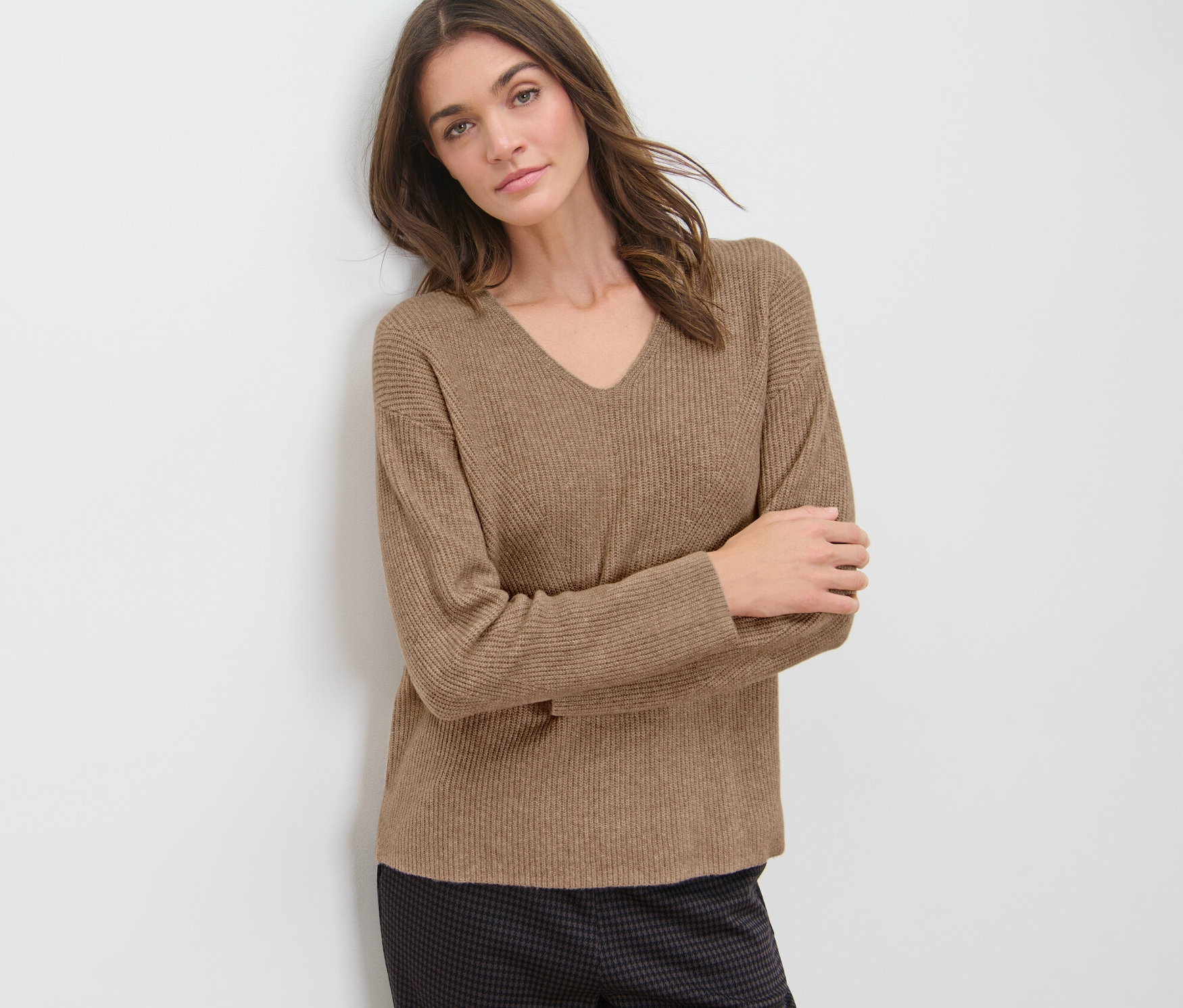 A falnak dőlő nő laza kötésű, tevebarna pulóvert visel.