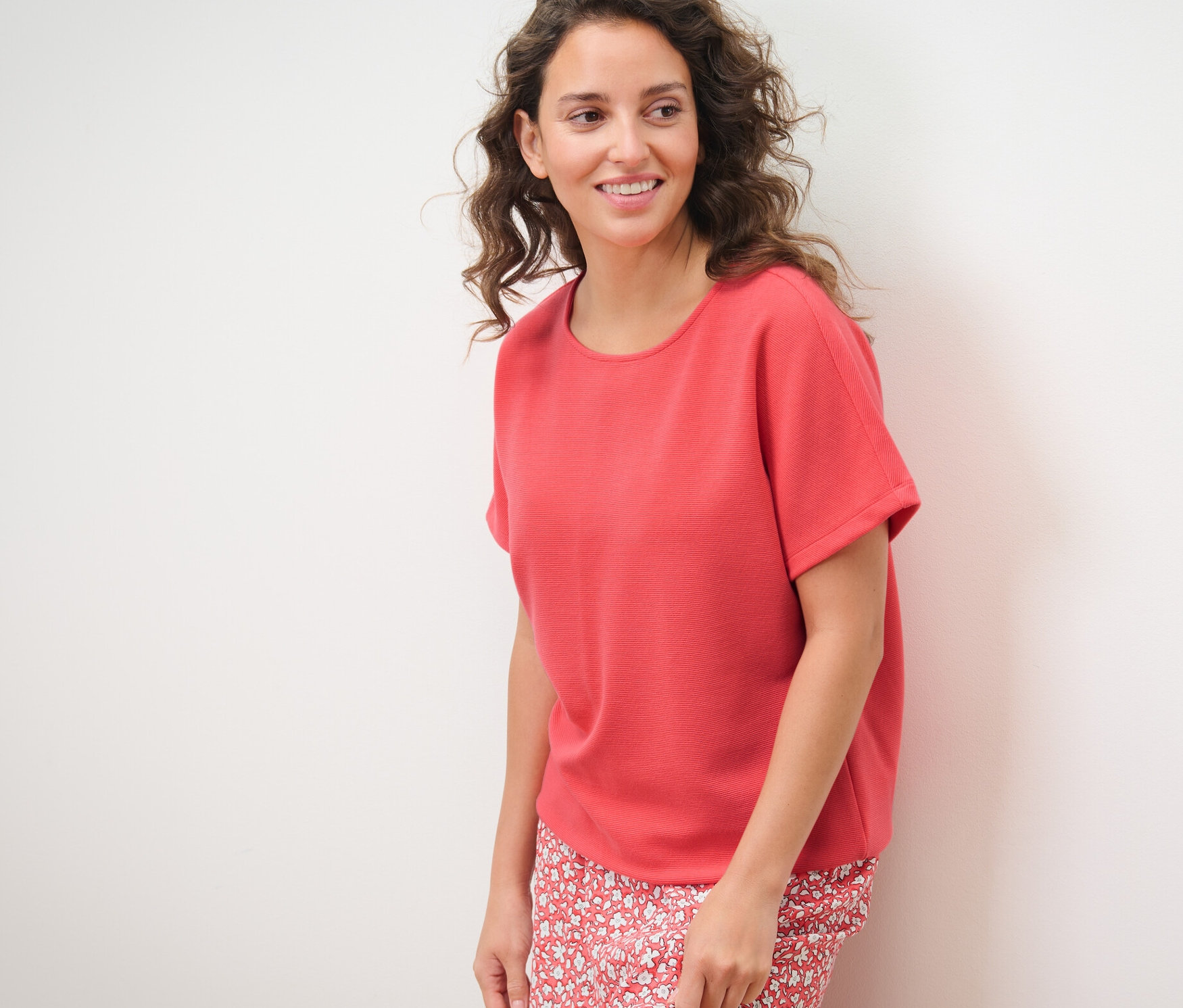 Nő pózol fehér háttér előtt, korall színű női pólóblúzt visel.