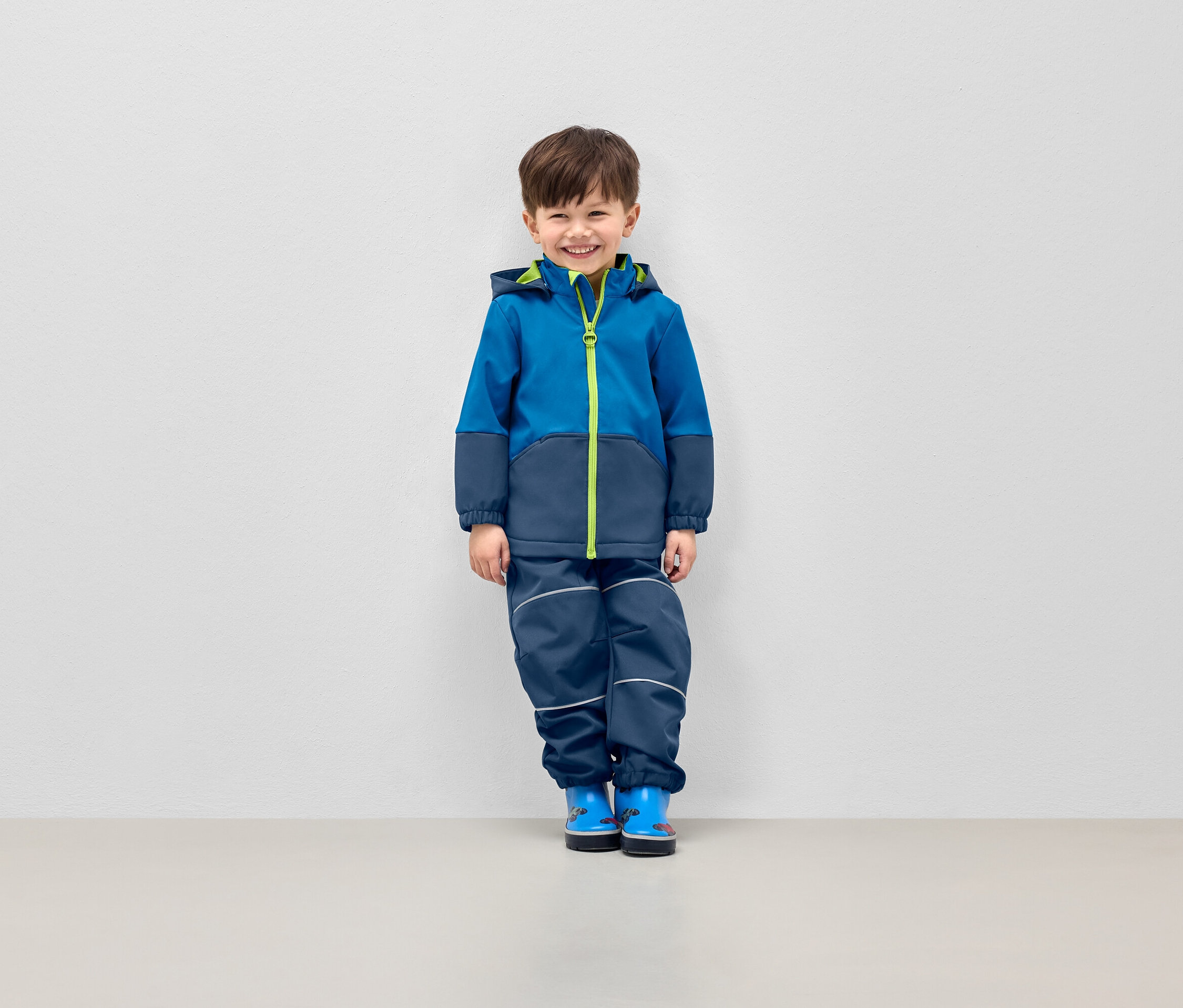 Mosolygó fiú pózol kék kapucnis kabátban és sötétkék nadrágban fényvisszaverő csíkokkal.