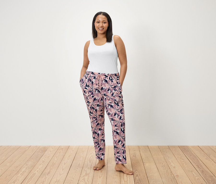 Egy nő áll virágmintás nadrágjának zsebében tartva a kezét. Fehér trikót visel.