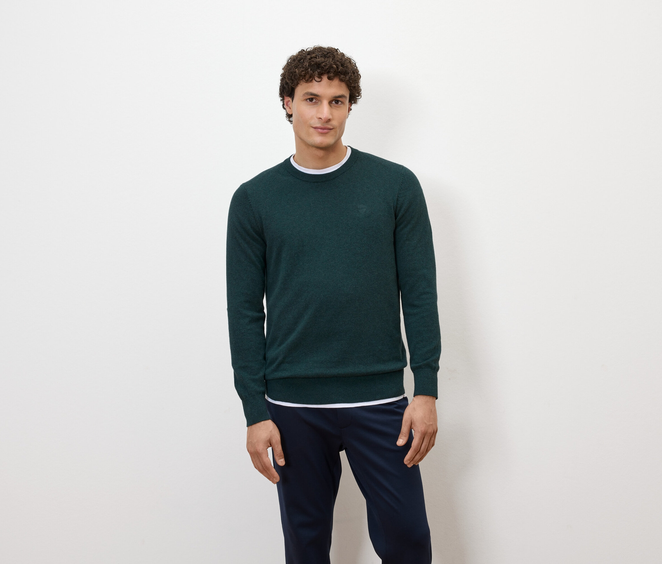 Egy férfi pózol fehér háttér előtt, sötétzöld pulóverben és sötétkék nadrágban.