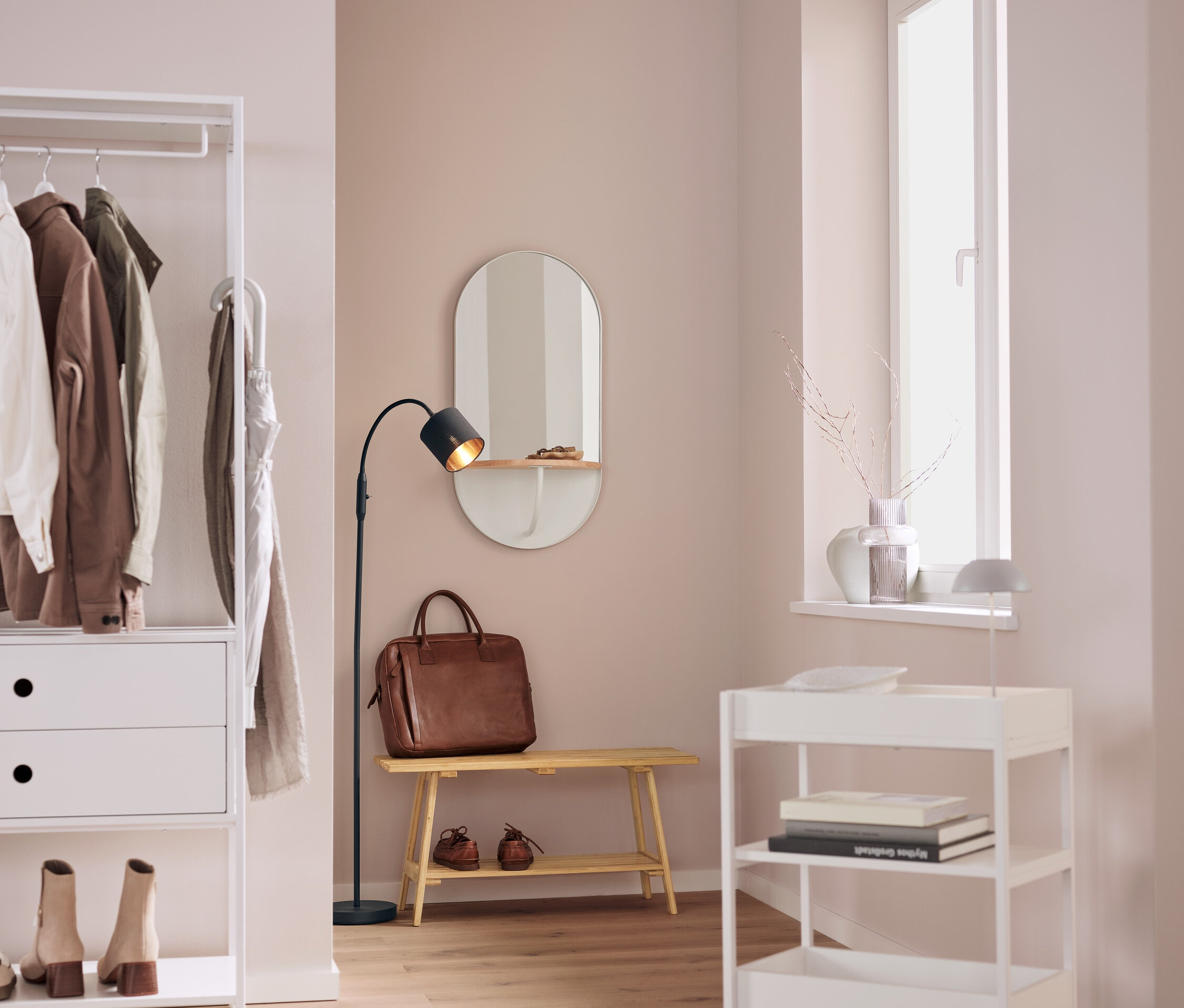 Egy belső térben egy fehér polc ruhákkal, egy ovális tükör, egy barna bőrtáska egy padon, egy fekete lámpa és egy fehér gurulós kocsi található.