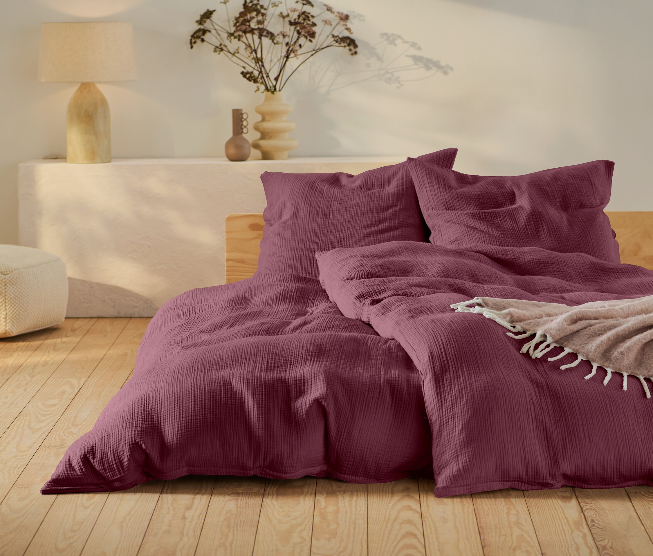 Bordó ágynemű fa keretes ágyon, bézs puff és egy lámpa egy fehér asztalon.