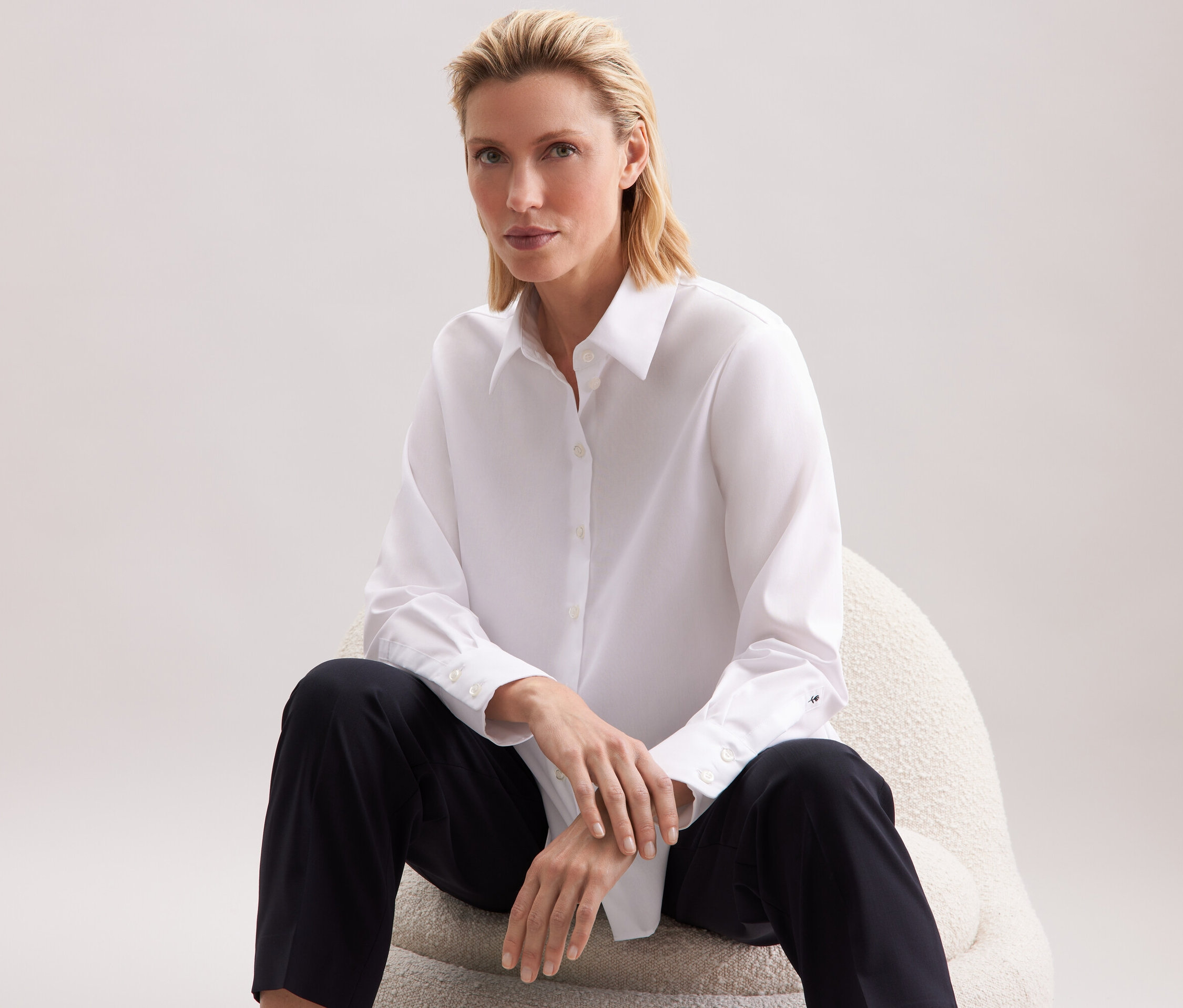 Nő ül egy széken, fehér inget és fekete nadrágot visel.