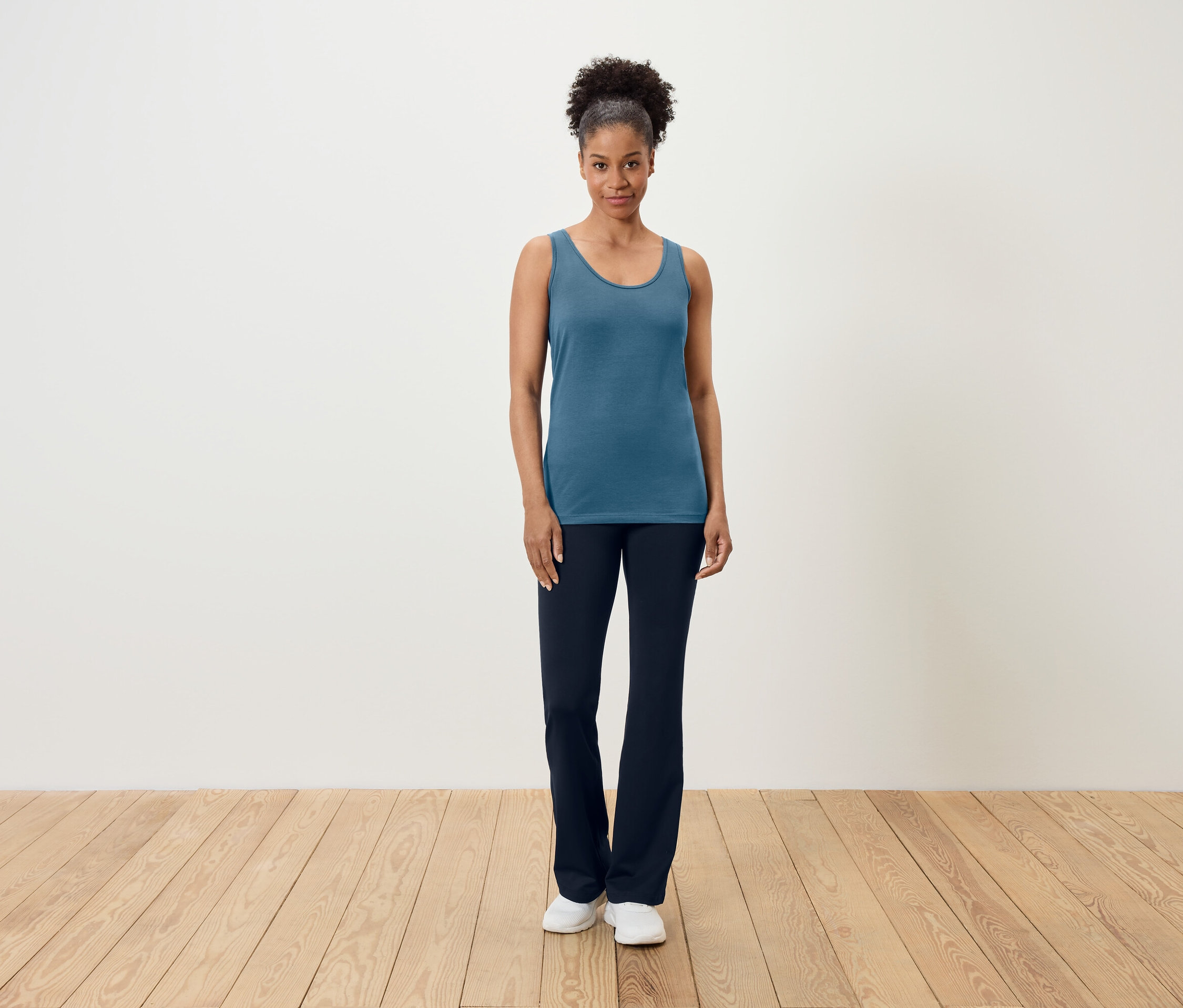 Egy nő áll egy fa padlón, kék trikót és sötétkék nadrágot visel.