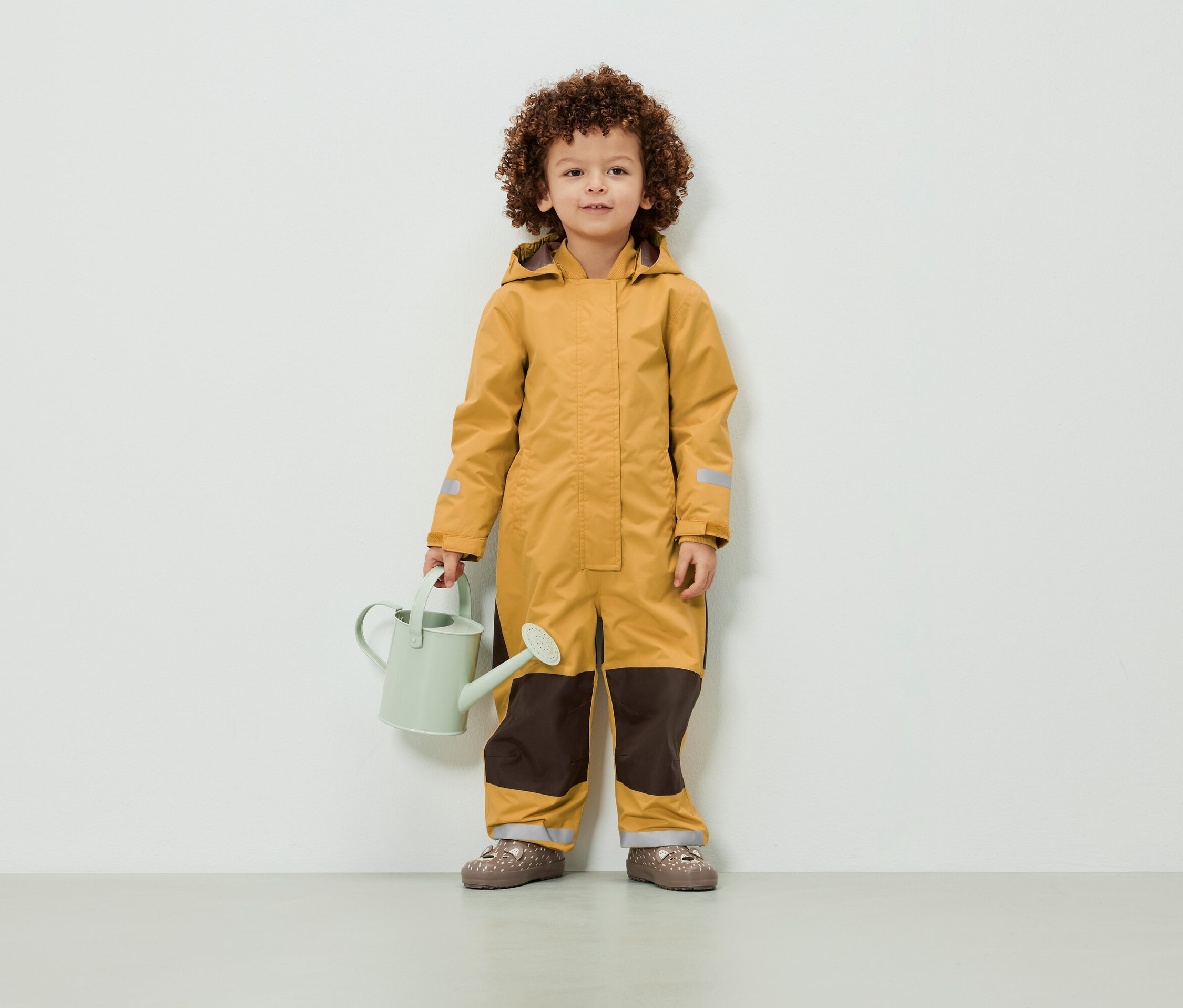 Egy göndör hajú gyermek sárga overallban áll, és egy világoszöld locsolókannát tart.