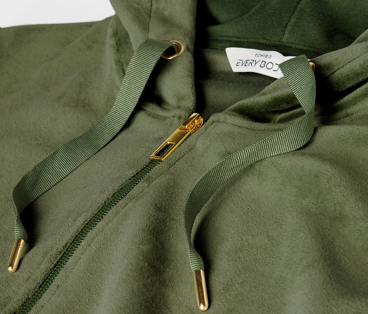 Egy zöld kapucnis pulóver részlete arany cipzárral és húzózsinórral.