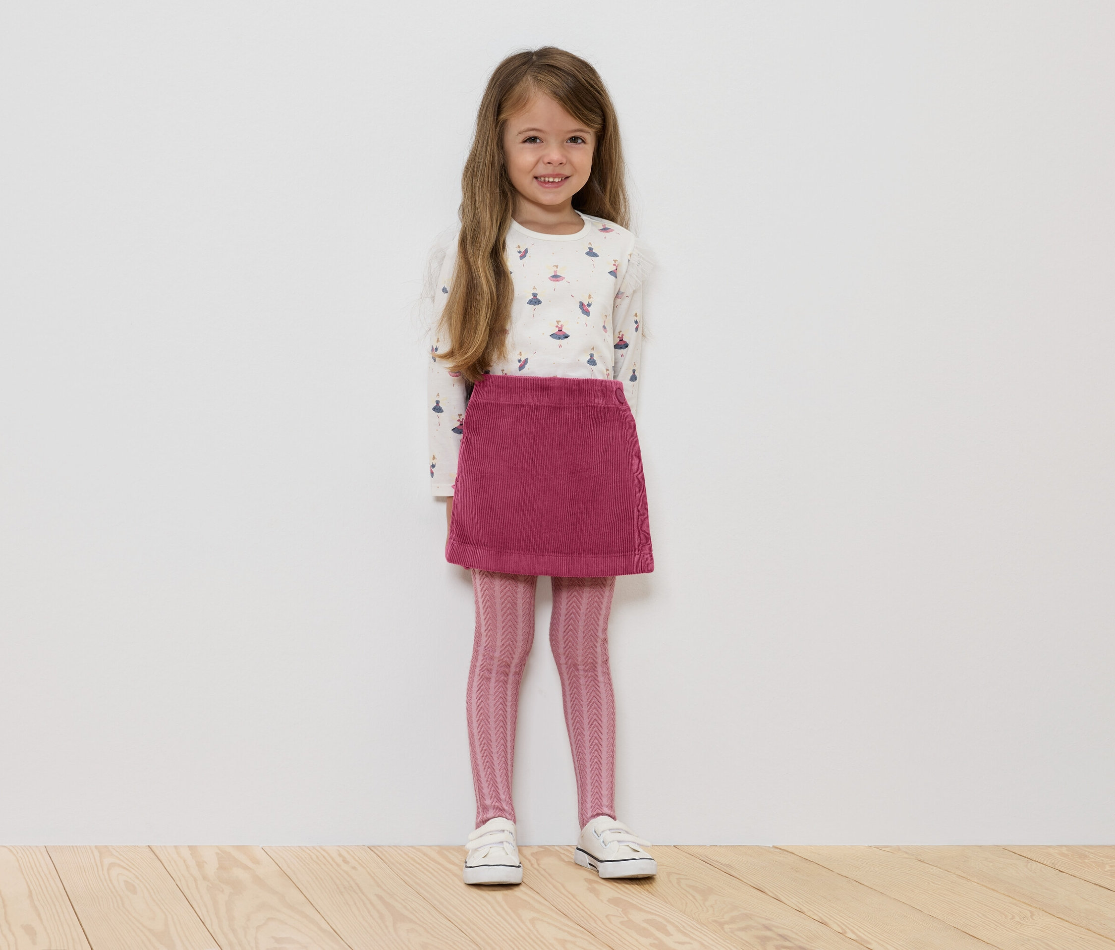 Egy kislány áll, fehér, hosszú ujjú, balerinás mintás pólót, rózsaszín kordbársony szoknyát, rózsaszín, mintás leggingset és fehér tornacipőt visel.