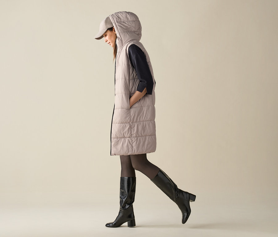 Egy nő sétál, steppelt, kapucnis mellényt, sapkát és térdcsizmát visel.