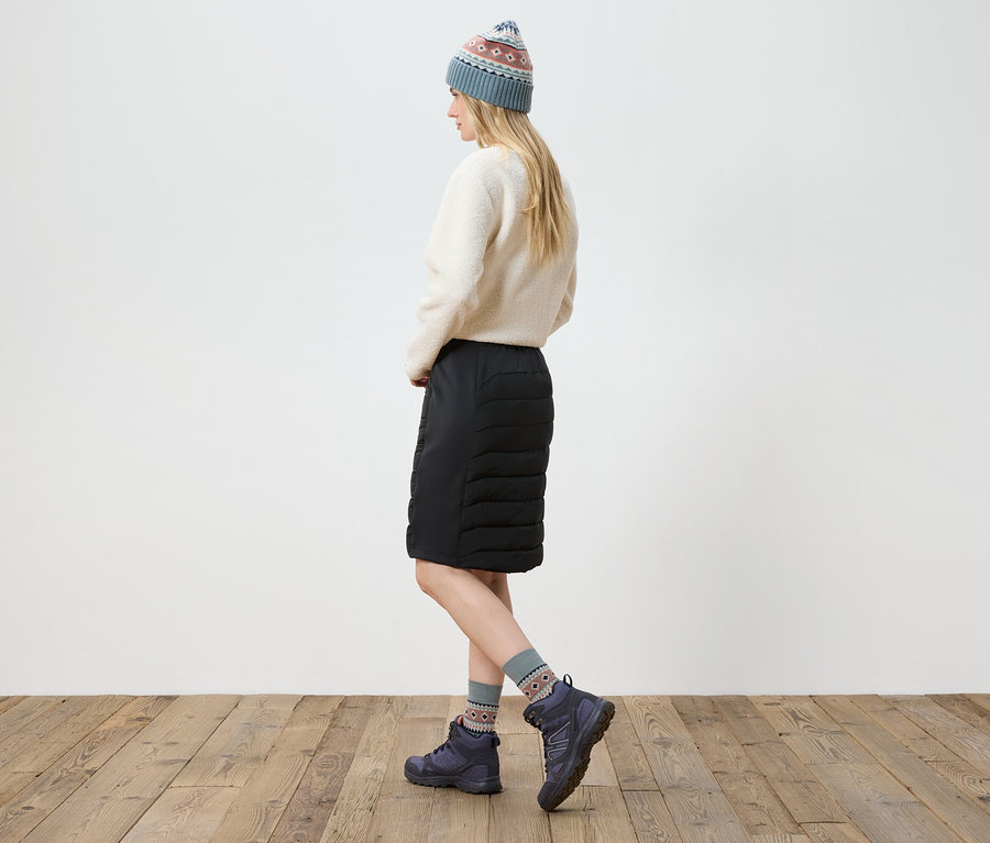 Nő pózol krémszínű pulóverben, fekete szoknyában, csizmában és kötött sapkában.