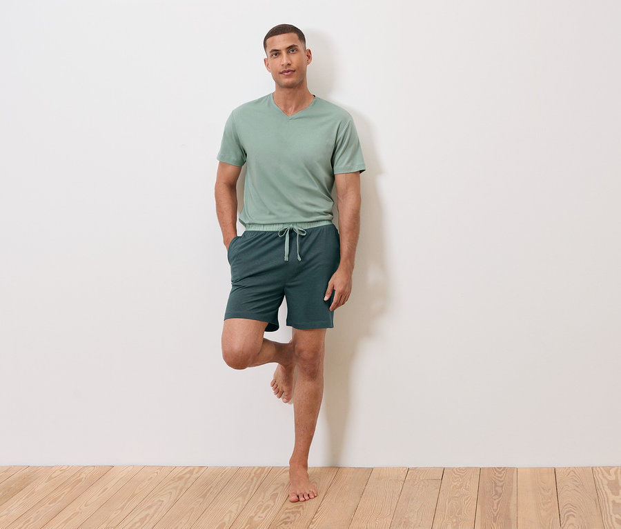 Férfi áll egy lábon a falnak támaszkodva, zöld pólót és rövidnadrágot visel.