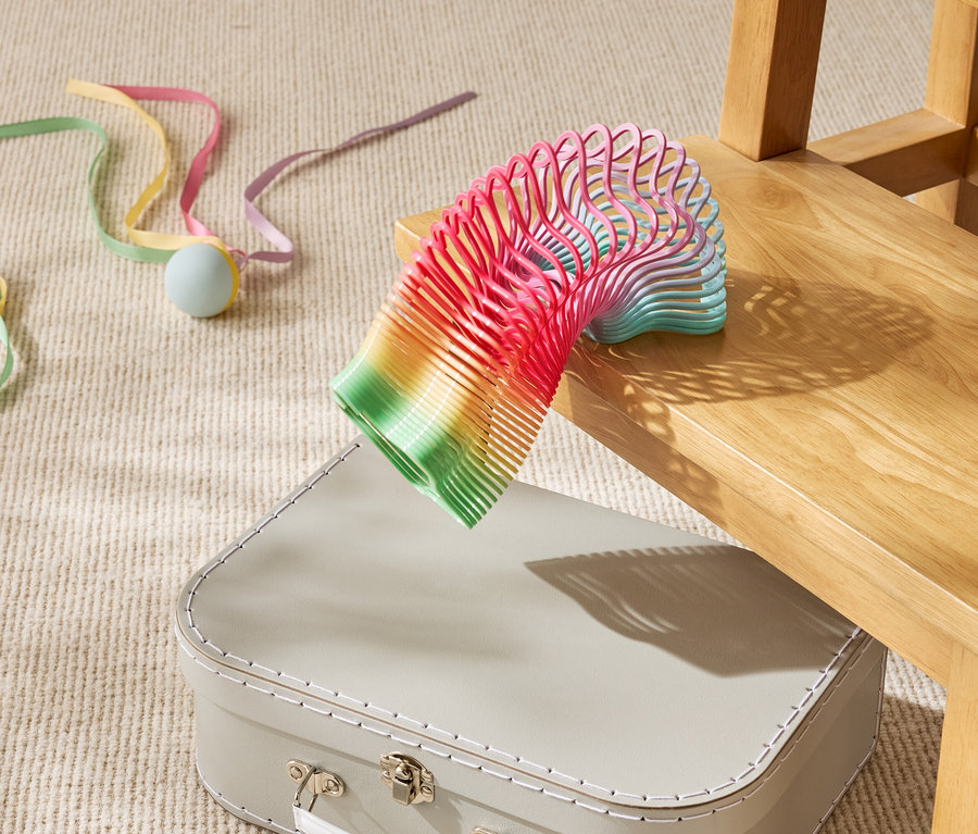 Egy szivárványszínű játék spirál részben egy bézs bőröndön fekszik egy fa szék alatt.