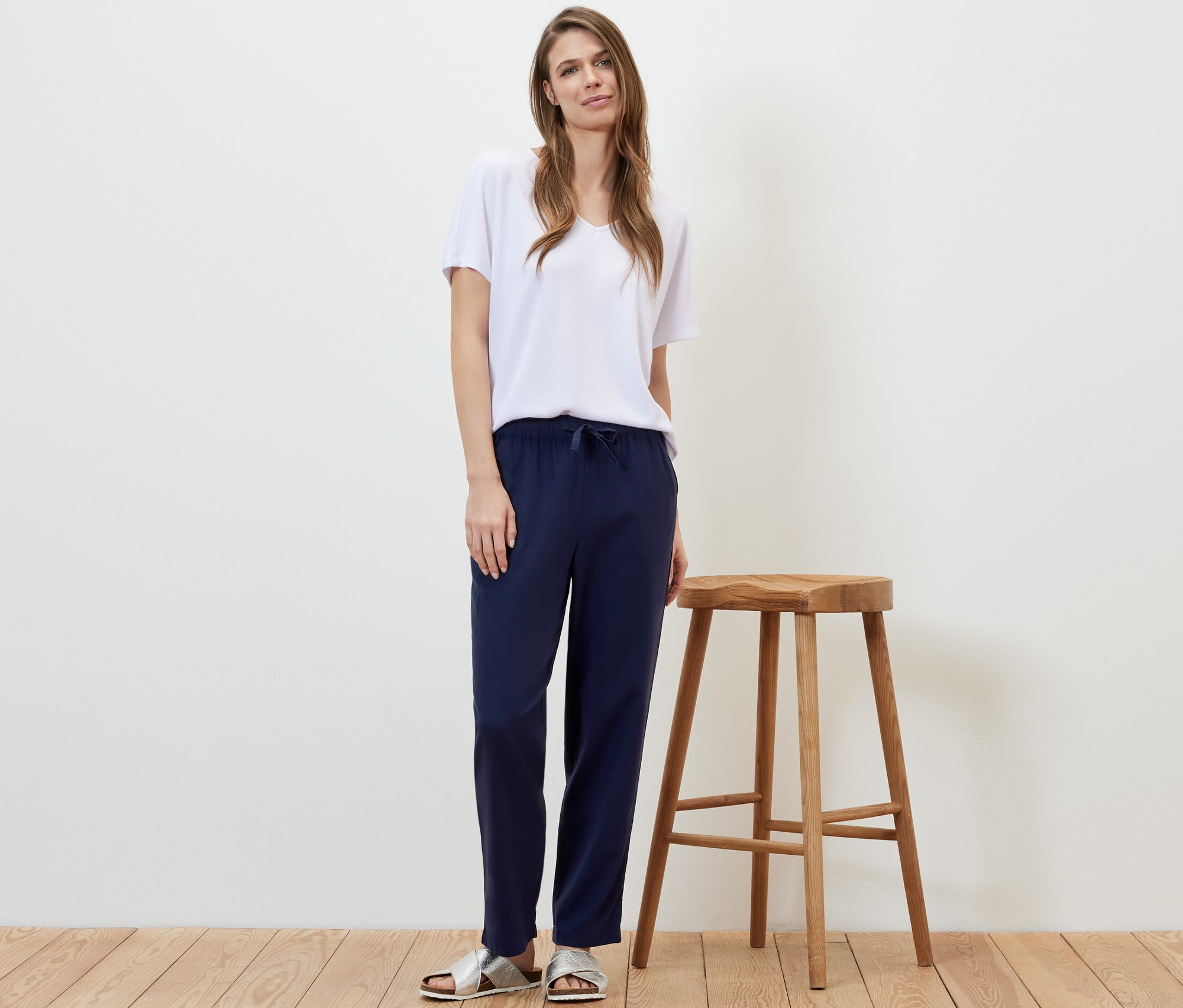 Egy nő egy fa zsámoly mellett áll, sötétkék vászonnadrágot és fehér pólót visel.