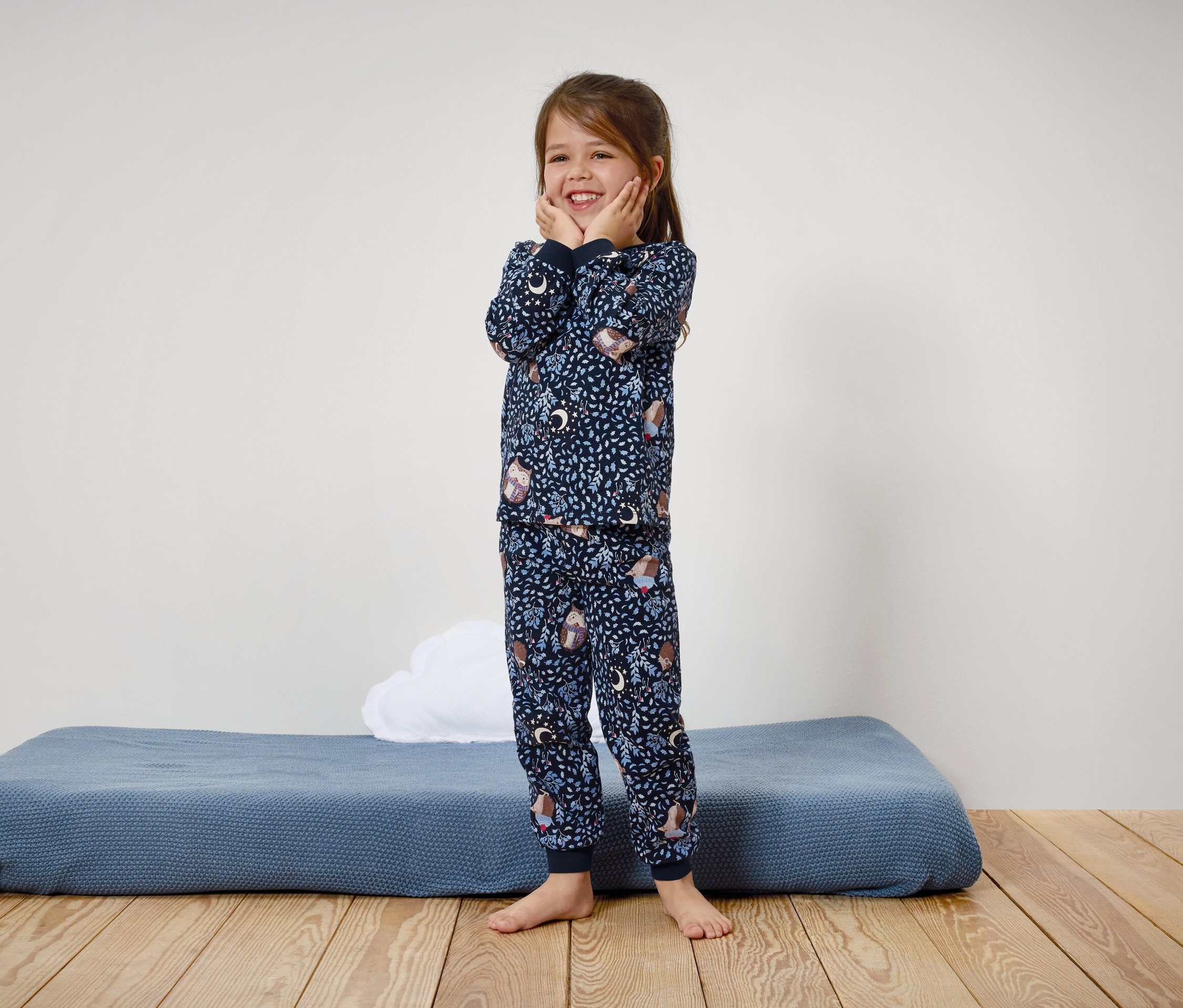 Egy mosolygós kislány kék, mintás pizsamában áll a fapadlón.