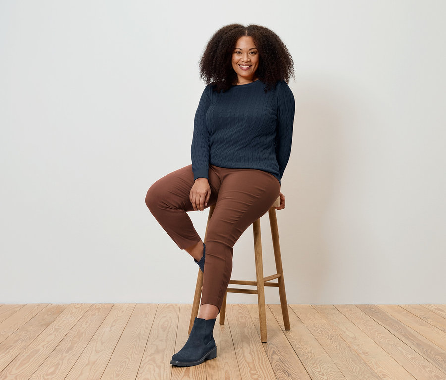 Egy nő ül egy fa székleten, sötétkék pulóvert, barna nadrágot és kék csizmát visel.