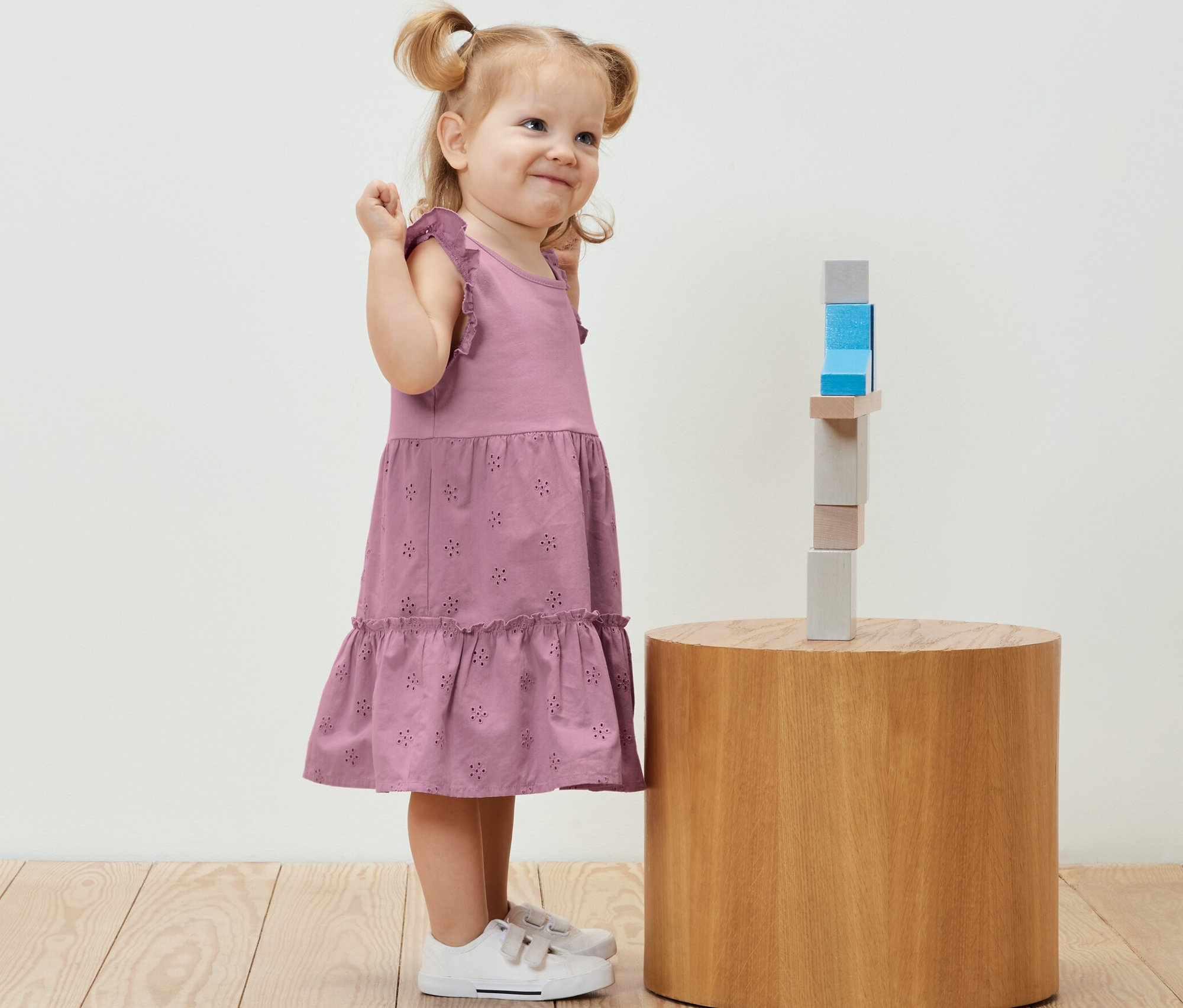 Mosolygó kislány gyerekszoknyában fa építőkockák mellett áll egy kerek asztalon.