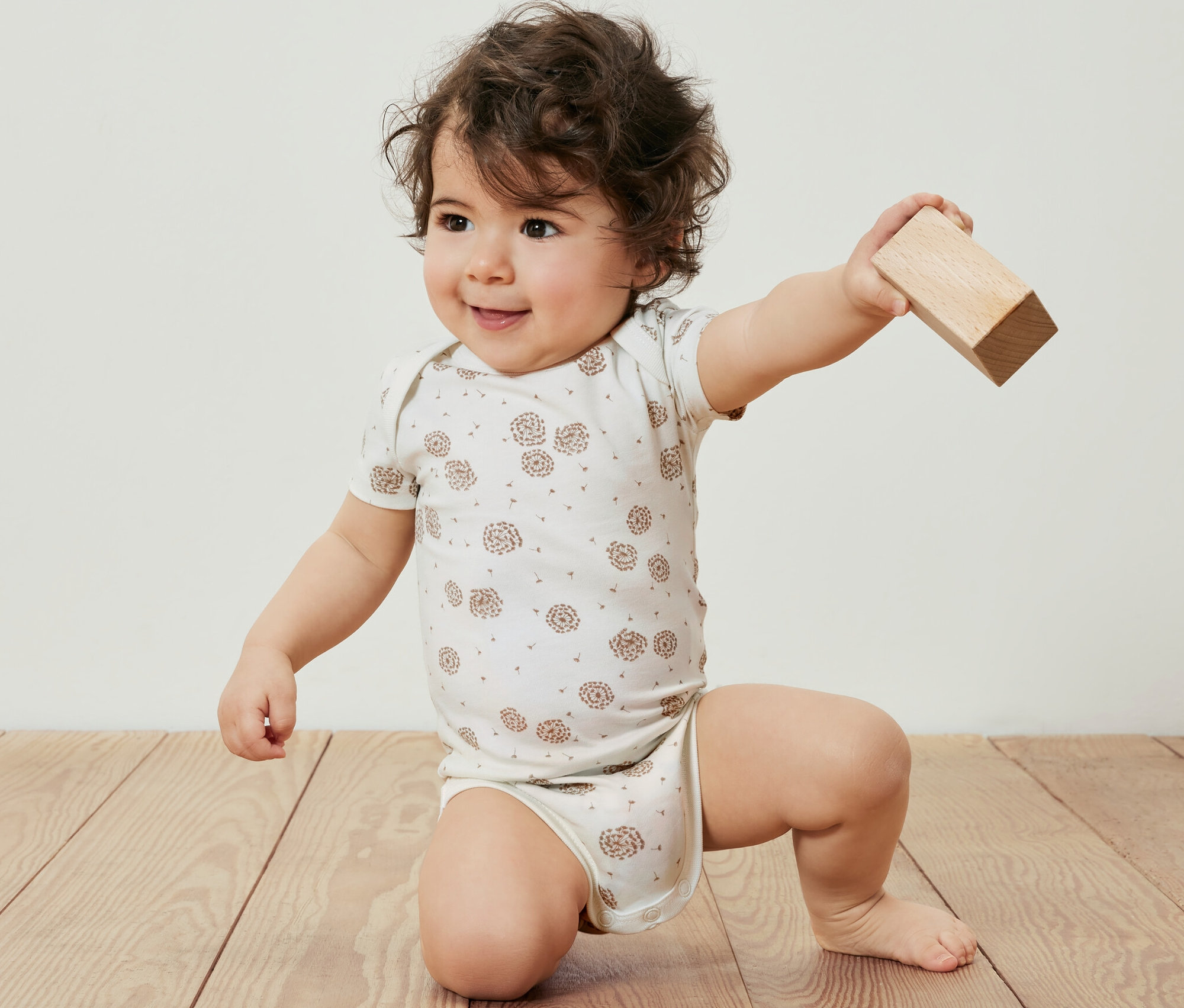 Egy kisgyerek az 5 darabos, állítható hosszúságú bodyban térdel a fapadlón, és egy fatömböt tart a kezében.