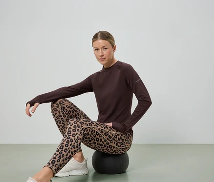 Nő ül egy fekete labdán, barna hosszú ujjú felsőt, leopárdmintás leggingset és fehér tornacipőt visel.