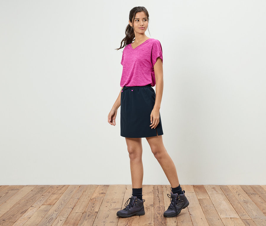 Egy nő áll egy fehér fal előtt, rózsaszín pólót, fekete szoknyát és túrabakancsot visel.